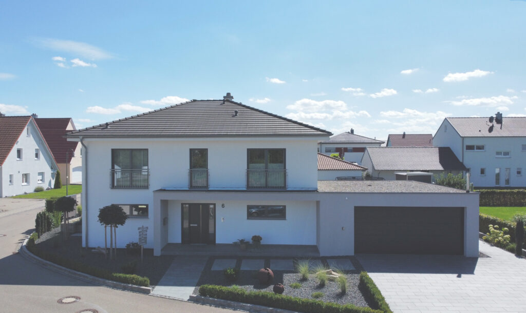 Modernes zweistöckiges weißes Haus mit grauem Dach, großen Fenstern und einer Doppelgarage. Kleiner begrünter Vorgarten mit Sträuchern und Steinweg. Andere ähnliche Häuser sind im Hintergrund unter blauem Himmel zu sehen.