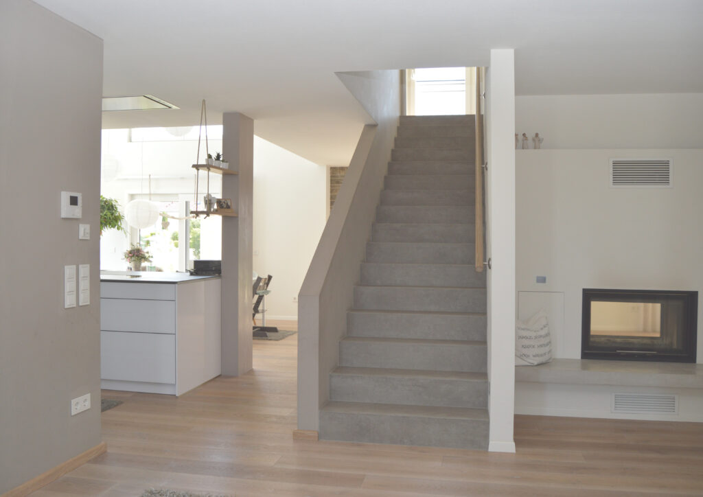 Das moderne, minimalistische Interieur verfügt über eine zentrale Treppe mit grauen Stufen, helle Holzböden, einen eingebauten Kamin auf der rechten Seite und einen offenen, hellen Küchenbereich auf der linken Seite mit Pflanzen und natürlichem Licht.