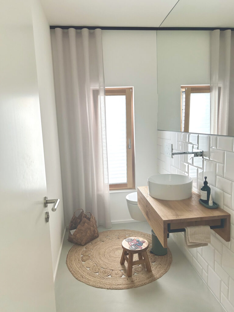Ein modernes Badezimmer mit einem runden gewebten Teppich, einem kleinen Holzhocker, einem schwimmenden Waschbecken aus Holz, einem Gefäßwaschbecken, einem Seifenspender, einem Fenster mit durchsichtigen Vorhängen und weiß gekachelten Wänden.