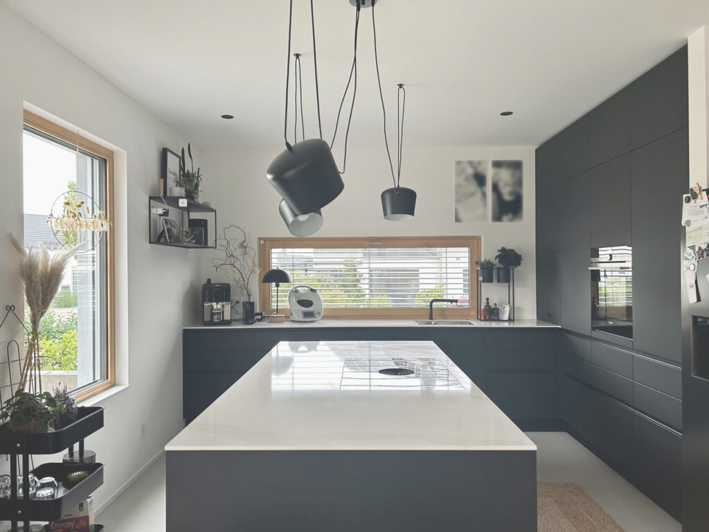 Moderne Küche mit schwarzen Schränken, einer großen weißen Insel, Hängelampen und einem Fenster mit Jalousien, das natürliches Licht hereinlässt. Pflanzen und Küchengeräte sind ordentlich auf Theken und Regalen angeordnet.