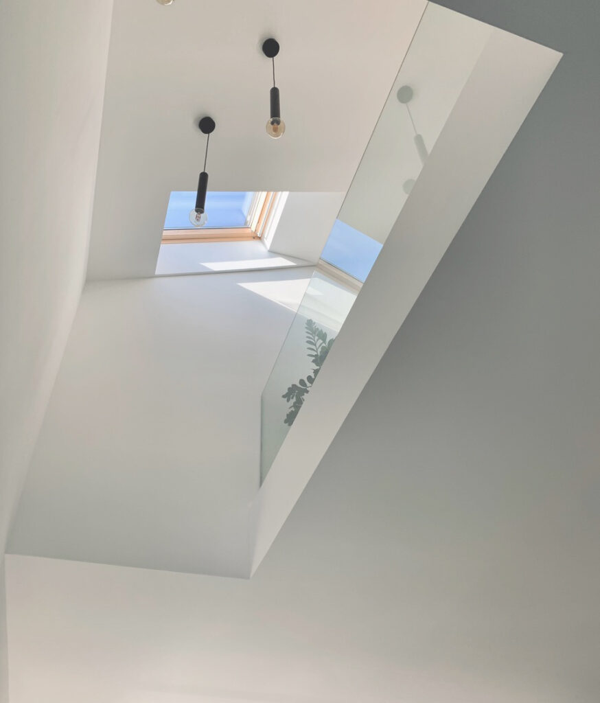Eine moderne, minimalistische Decke mit einem Oberlicht, in dem der blaue Himmel zu sehen ist, zwei hängenden Glühbirnen und einem Glasgeländer, das das Licht reflektiert. Auf dem Glas spiegelt sich auch eine Blattpflanze.