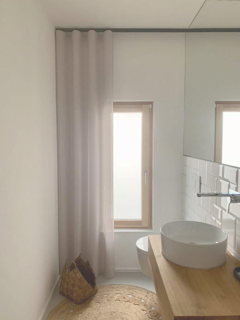 Minimalistisches Badezimmer mit einem runden Waschbecken auf einem Holztisch, einem kleinen Fenster mit weißen Vorhängen, einem Spiegel, weiß gefliesten Wänden, einem geflochtenen Korb und einem beigen Teppich auf dem Boden.