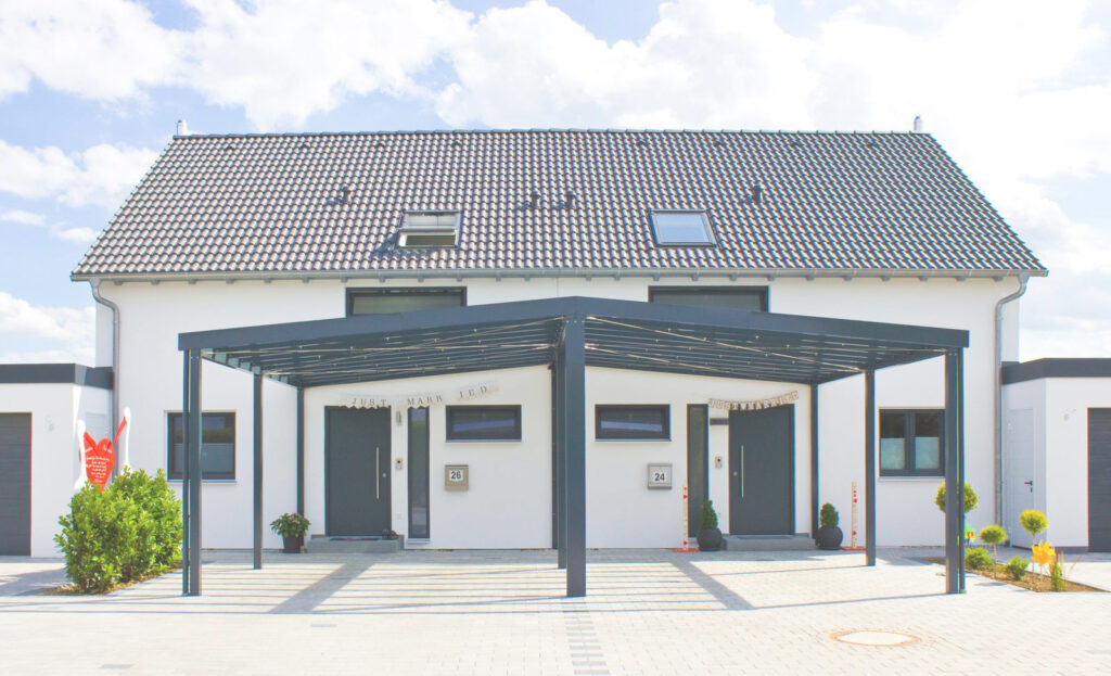Ein modernes weißes Doppelhaus mit dunklem Ziegeldach und Oberlichtern, mit einem gemeinsamen Metallcarport an der Vorderseite. Die Eingangstüren liegen nebeneinander, die Hausnummern 23 und 24 sind sichtbar. Die Einfahrt ist gepflastert, und an den Seiten befinden sich kleine Büsche.