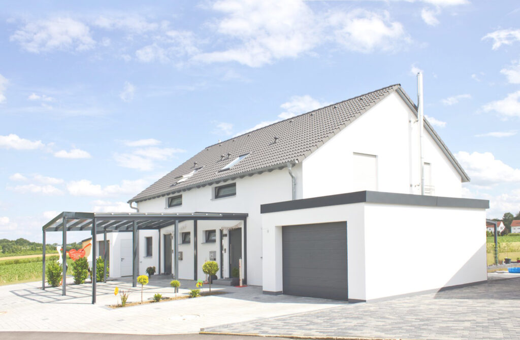Ein modernes weißes zweistöckiges Haus mit grauem Dach, angebauter Garage und gläsernem Carport steht auf einer gepflasterten Auffahrt, umgeben von kleinen Landschaftsbüschen und offenen Feldern unter einem teilweise bewölkten Himmel.