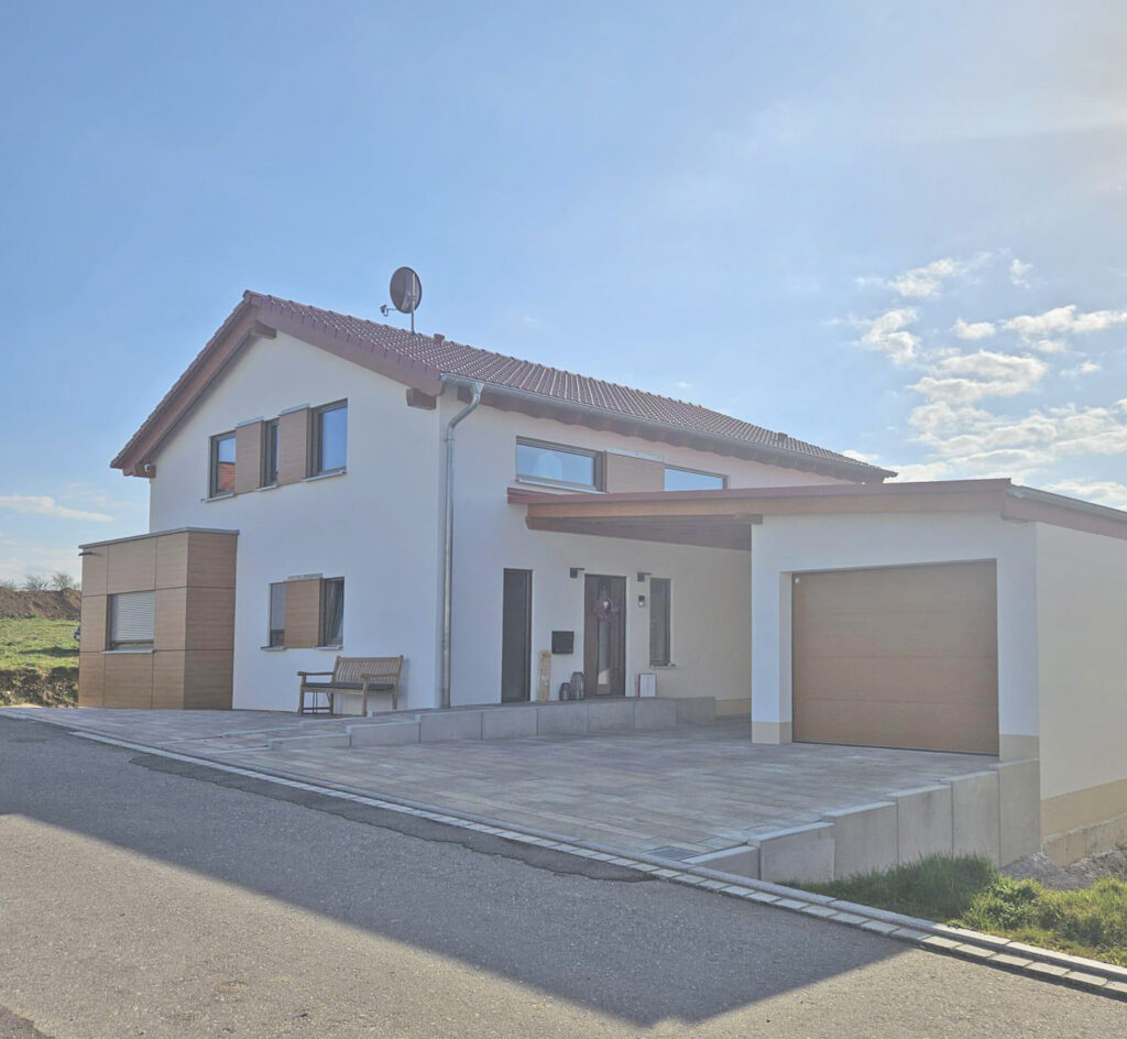 Ein modernes, zweistöckiges, weißes Haus mit rotem Ziegeldach, angebauter Garage und einer kleinen Veranda, direkt an einer asphaltierten Straße unter einem klaren, sonnigen Himmel gelegen.