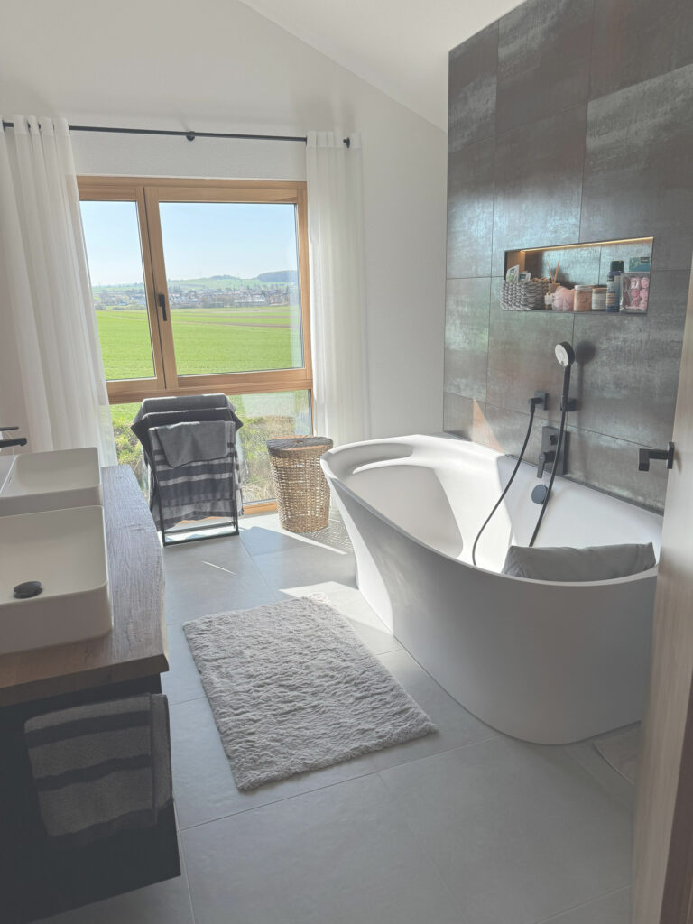 Modernes Badezimmer mit freistehender weißer Badewanne, Doppelwaschbecken auf einem hölzernen Waschtisch, großem Fenster mit weißen Vorhängen, Handtuchhalter, Weidenkorb und Regalen mit Toilettenartikeln vor einer gefliesten Wand. Der Raum ist von natürlichem Licht durchflutet.