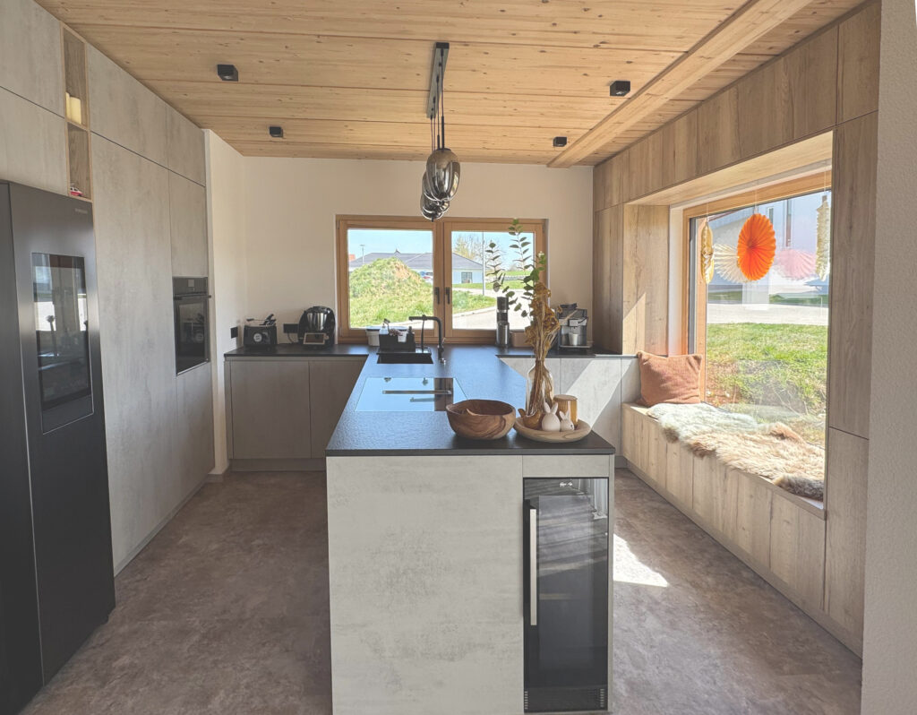 Moderne Küche mit hellem Holz und grauen Oberflächen, einer zentralen Insel, Einbaugeräten, einer Kaffeemaschine und einem Fensterplatz mit Kissen und einer Decke mit Blick auf einen grasbewachsenen Hof vor großen Fenstern.