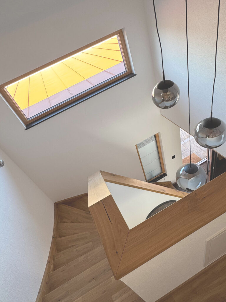 Eine moderne Holztreppe mit schrägen Stufen führt hinunter zu einem gefliesten Boden. Es hängen drei kugelförmige Pendelleuchten, und ein großes Fenster sorgt für helles Tageslicht von oben.