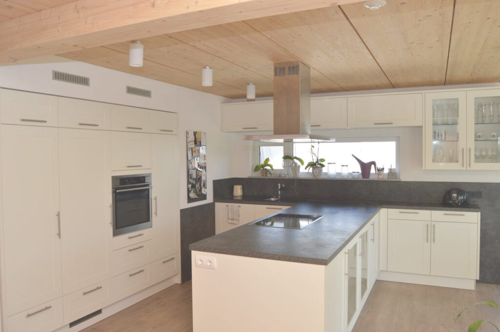 Moderne Küche mit weißen Schränken, Einbaubackofen, Insel mit Spüle, grauer Arbeitsplatte, Topfpflanzen am Fenster und Holzdecke mit Einbaubeleuchtung. Auf der linken Seite ist ein Kühlschrank mit Fotos zu sehen.