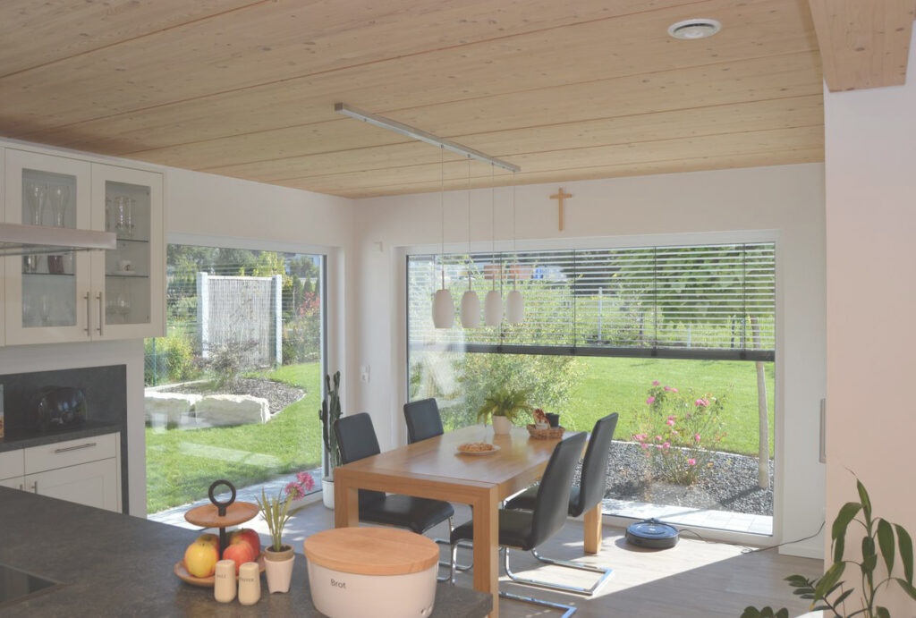 Ein moderner Essbereich mit einem Holztisch, sechs schwarzen Stühlen, großen Fenstern mit Blick auf einen Garten und Pendelleuchten über dem Tisch. Auf der Theke im Vordergrund stehen Obst und Küchengeräte.