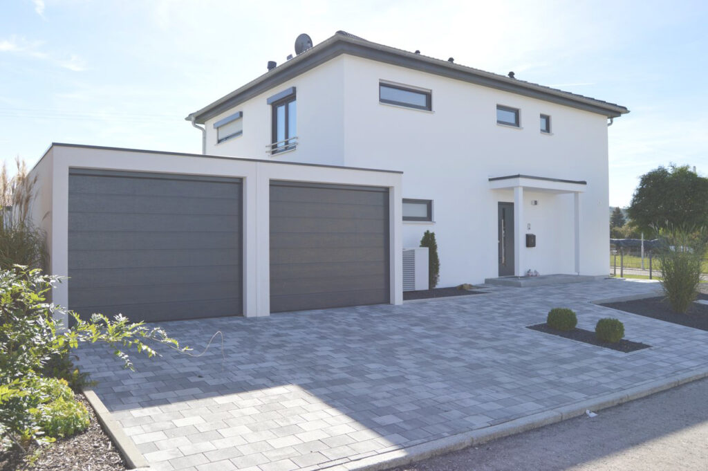Modernes weißes zweistöckiges Haus mit Flachdach, großen Fenstern und einer Doppelgarage. Die Einfahrt ist mit grauen Ziegeln gepflastert, und vor dem Gebäude ist eine minimale Bepflanzung mit kleinen Sträuchern zu sehen.