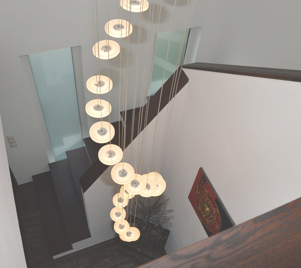 Ein modernes Treppenhaus mit Stufen und Geländern aus dunklem Holz und einem vertikalen Kronleuchter mit runden, spiralförmig aufgehängten Leuchten. Große Milchglasfenster und ein dekoratives rotes Wandkunstwerk sind ebenfalls zu sehen.