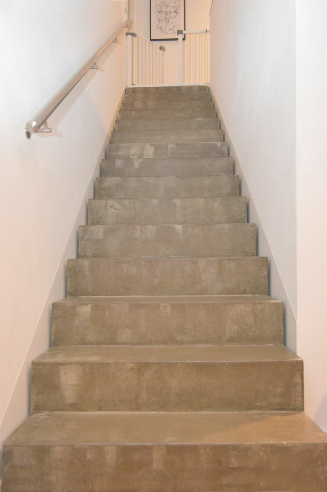 Eine Betontreppe mit einem Metallhandlauf auf der linken Seite, die zu einem weißen Kindergitter am oberen Ende führt. An der Wand am Ende der Treppe hängt eine gerahmte schwarz-weiße Strichzeichnung.