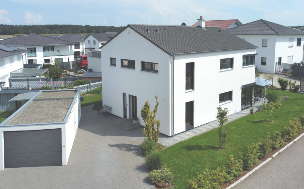 Ein modernes zweistöckiges weißes Haus mit grauem Dach, großen Fenstern und einer freistehenden Garage, umgeben von grünem Rasen, kleinen Bäumen und Nachbarhäusern in einer Vorstadt.